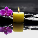 burning yellow candle with purple orchid on black rocks and water