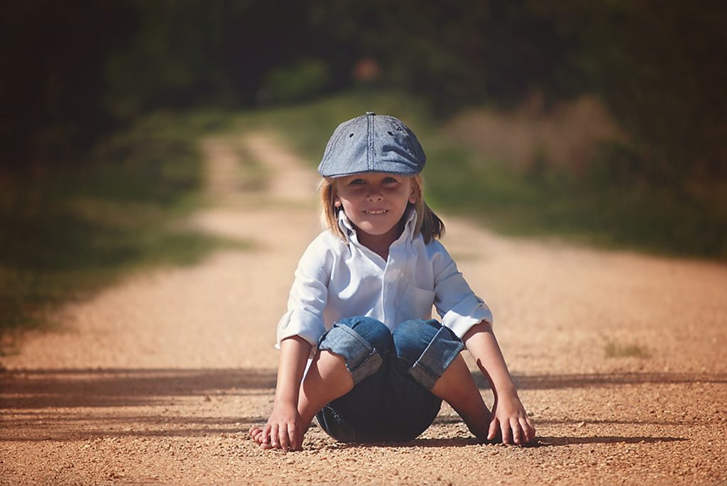 Little boy, wearing a cap, sitting in the middle of a dirt road by Lisa Runnels