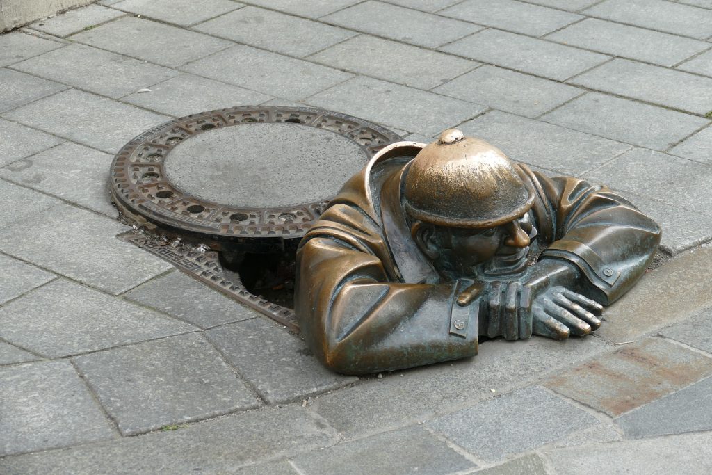 A photo of a sculpture of the top portion of a worker coming out of a manhole, leaning on the sidewalk with chin resting on his crossed arms. Image by Falco on Pixabay